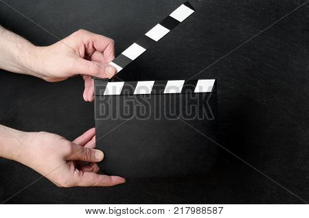 A blank clapperboard held open as if about to snap shut between the hands of a male operative with black chalkboard background.