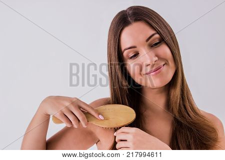 She loves her hair. Cropped image of beautiful brunette girl combing hair and smiling while isolated on white background. Young woman wrapped in towel with comb in hand. Hair care and treatment