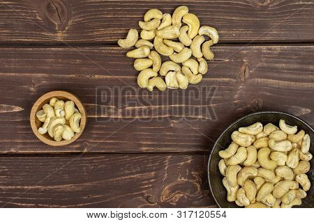 Lot Of Whole Unsalted Beige Cashew In Tiny Wooden Bowl In Dark Ceramic Bowl Flatlay On Brown Wood