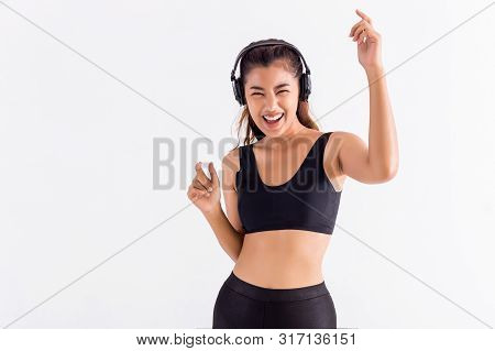 Happy Asian Woman In Black Sport Bra Listening To Music And Dancing With Raised Hands On White Backg