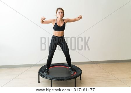 Attractive Young Woman In Sportswear Jumping On Mini Trampoline Against White Wall During High Inten