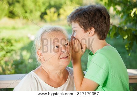 Boy whispering to grandma in summer outdoors garden, grandkid and grandmother lalking.