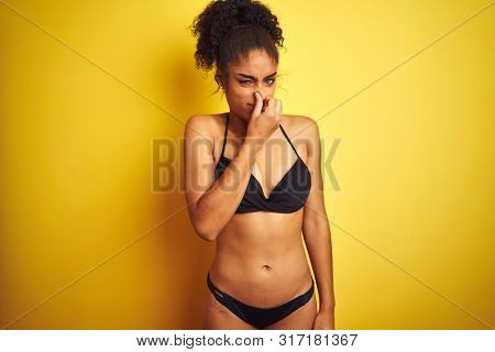 African american woman on vacation wearing bikini standing over isolated yellow background smelling something stinky and disgusting, intolerable smell, holding breath with fingers on nose. Bad smells 