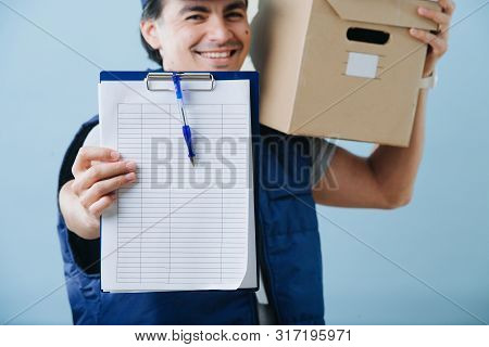 Delivery Man In Uniform Is Holding Parcel Cardboard Box On His Shoulder Over Blue Background. He Is 