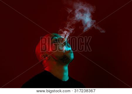 Close-up Studio Shot Of A Young Bald Guy Vaping, Blowing Out A Cloud Of Smoke On Red Background.