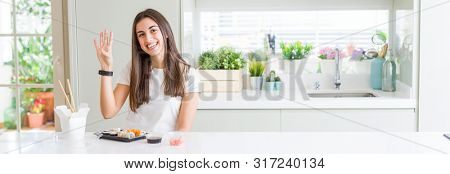 Wide angle picture of beautiful young woman eating asian sushi from delivery showing and pointing up with fingers number four while smiling confident and happy.