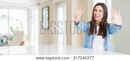 Wide angle picture of beautiful young woman sitting on white table at home showing and pointing up with fingers number eight while smiling confident and happy.