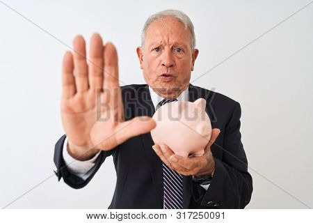 Senior grey-haired businessman holding piggy bank standing over isolated white background with open hand doing stop sign with serious and confident expression, defense gesture