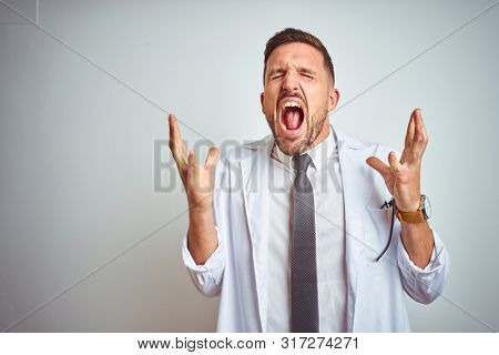 Young handsome doctor man wearing white profressional coat over isolated background celebrating mad and crazy for success with arms raised and closed eyes screaming excited. Winner concept