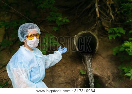 Female Ecologist Or Epidemiologist In A Protective Gown And Mask Points To A Sewer Coming Out Of The
