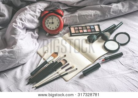 Make Up Cosmetic And Brushes, Alarm Clock And Blank Page Notepad On The Blanket On The Bed Backgroun