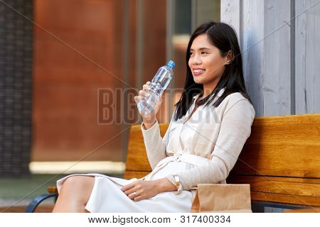 lifestyle and people concept - happy smiling young asian woman drinking water flom plastic bottle sitting on wooden city bench