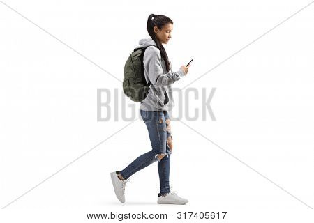 Full length profile shot of a female student with a backpack walking and looking into a mobile phone isolated on white background