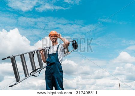 Cheerful Repairman Holding Ladder And Smiling Against Blue Sky With Clouds