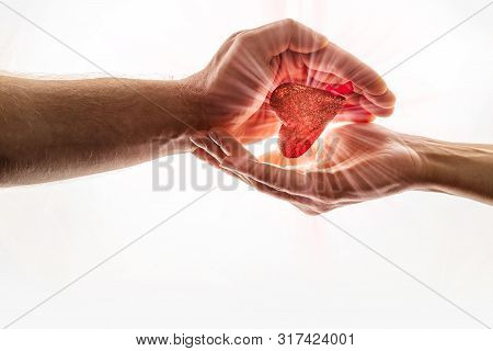 Female And Male Hands Are Keeping Red Heart Together. Heart With Brightly Ray Of Sunshine Is Between