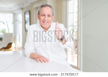 Handsome senior man at home doing happy thumbs up gesture with hand. Approving expression looking at the camera with showing success.