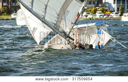 A Girl And Boy In The Water Climbing Back Into A Capsized Racing Sailboat. Teamwork By Junior Sailor