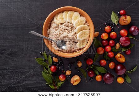 Image Of Oatmeal With Banana, Alich, Pluns, Bluberries, Pits And Green Leaves On Dark Wooden Surface