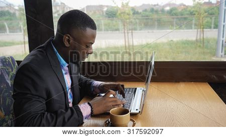 Afro American Businessman Is Typing A Message On Laptop Sitting In Summer Cafe Near Window. Black Ma