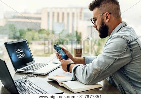 Hipster Man Sits In Cafe, Uses Smartphone, Works On Two Laptops With Inscription On Screen- Cloud Li