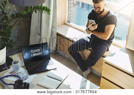 Hipster Man Sits Home On Windowsill, Reads Text Messages On Smartphone. On Table Laptop With Inscrip