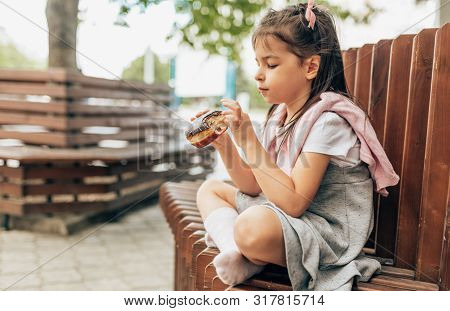Cute Little Girl Student Eating An Donut Sitting On The Bench Outdoor. Adorable Child Pupil Have A S