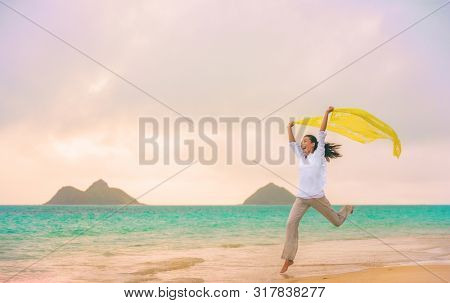 Happy woman jumping of joy and freedom running with yellow scarf in wind on Hawaii beach, Lanikai, Oahu, USA. Summer vacation fun free funny girl.