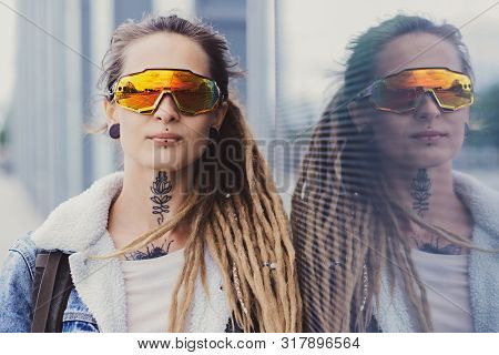 Beautiful Styilish Girl In Big Toned Glasses, Tattoo On The Neck And Piercing Is Posing Near Mirroed