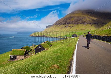 Young Skater Riding A Skateboard Through The Coastal Village Of Bour On The Faroe Islands Surrounded