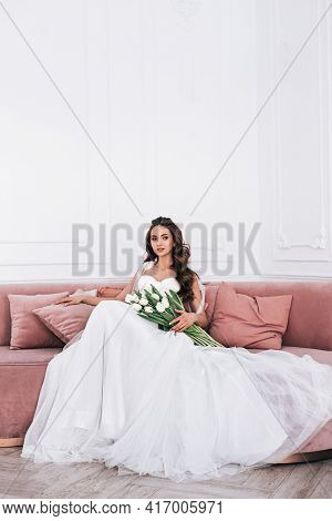 Bride In Beautiful Dress Holding Wedding Bouquet And Sitting On Sofa Indoors
