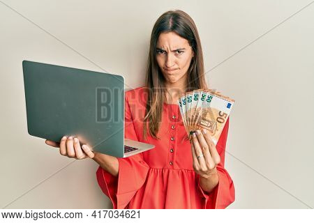 Young beautiful woman holding laptop and 50 euros skeptic and nervous, frowning upset because of problem. negative person. 