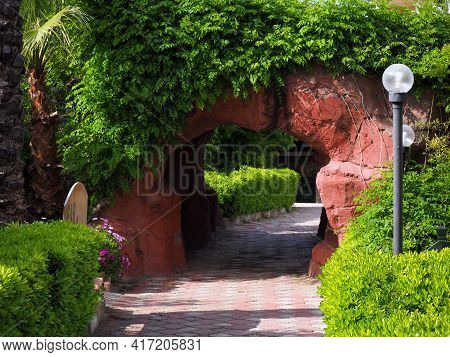 A Small Arched Tunnel From Above Is Overgrown With Bindweed. There Is A Lantern Near The Stone Arch,