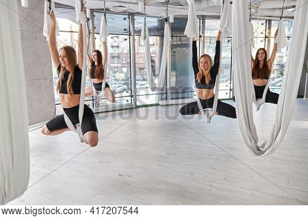 Group Of Female Are Doing Fly Yoga Exercises In Fitness Training White Gym Loft Classroom. Intense F