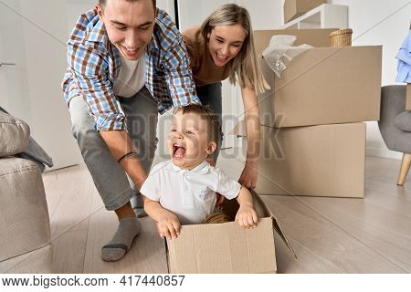Happy Parents Having Fun Playing With Cute Toddler Kid Son Riding In Box On Moving Day. Excited Youn