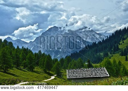 Alpine Hut On Pasture In Front Of Wood And Mountains With Clouds
