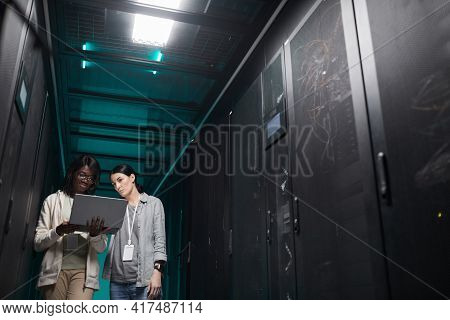 Low Angle Portrait Of Two Young Women Using Laptop In Server Room While Setting Up Supercomputer Net