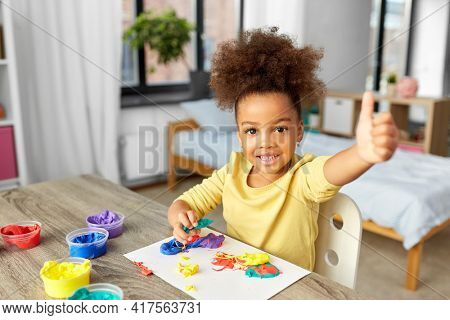 childhood, creativity and hobby concept - little african american girl playing with modeling clay and showing thumbs up at home
