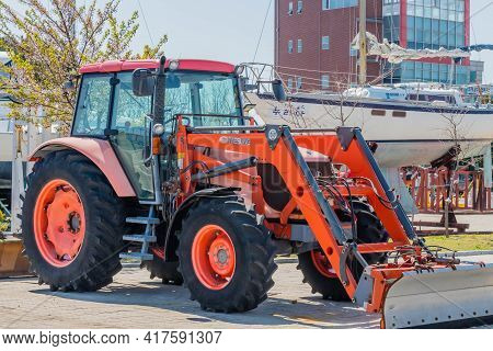 Red Tractor Parked At Marina In Front Of Sailboat.