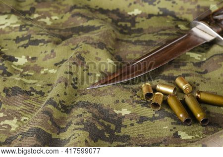 A Knife Blade And Some Random Casings On Camouflage Fabric. Knife Without Sheath And Group Of Pistol
