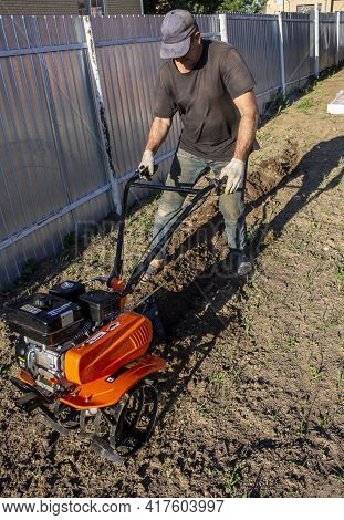 Male In The Spring Plows The Earth In Working Clothes With A Motoblock, Walk-behind Tractor