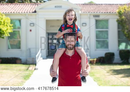 American Father And Son Walking Trough School Park. Man And Excited Amazed Kid Piggyback Near School
