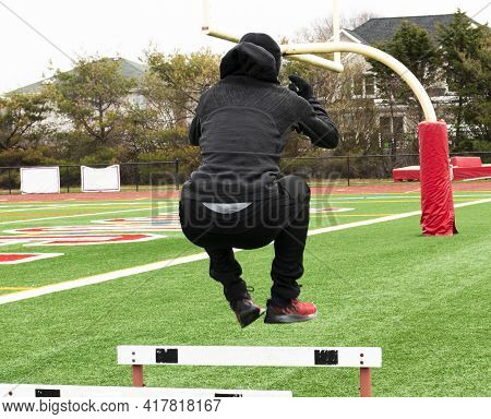 Rear View Of A Track And Field Athlete Jumping Over Hurdles On A Green Turf Field Wearing All Black.