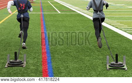 Rear View Of Two High School Male Track Runners Pulling Sleds With Weights On A Green Turf Field Dur