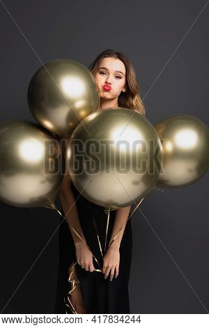Young Woman In Black Slip Dress Holding Golden Balloons And Pouting Lips On Grey.
