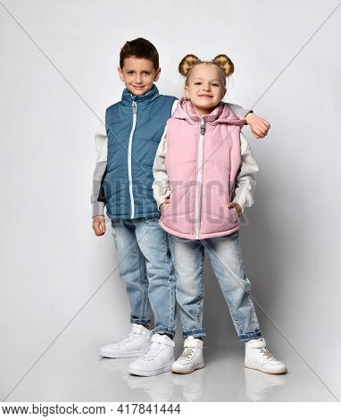Studio Shot Of Little Brother With Sister In Warm Outerwear Blue And Pink Vests, Jeans, Sneakers And
