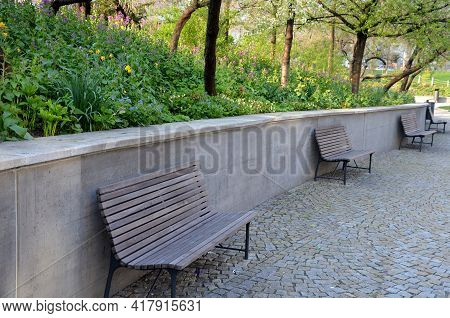 Bench At A Retaining Wall In A Park Of Black Bushes Early Spring Perennials On A High Flowerbed Path