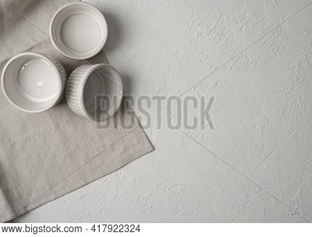 Flat Lay Of Three Empty White Ceramic Ramekins Stacked Against White Background, Served Linen Napkin