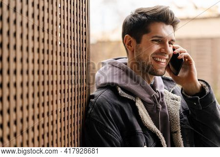 Smiling young white man in casual cothes using mobile phone outdoors in the city, leaning on a wall, talking