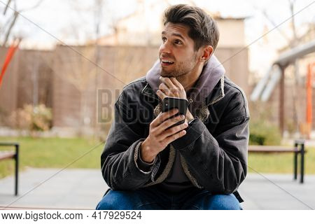 Smiling young white man in casual cothes using mobile phone outdoors in the city, sitting on a bench