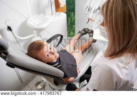 Boy Sitting In Dental Chair And Looking At Camera. Woman Dentist In Medical Mask Looking At Cute Chi